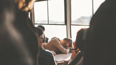 Woman on ferry with head resting in arms on small table with crowd of people around